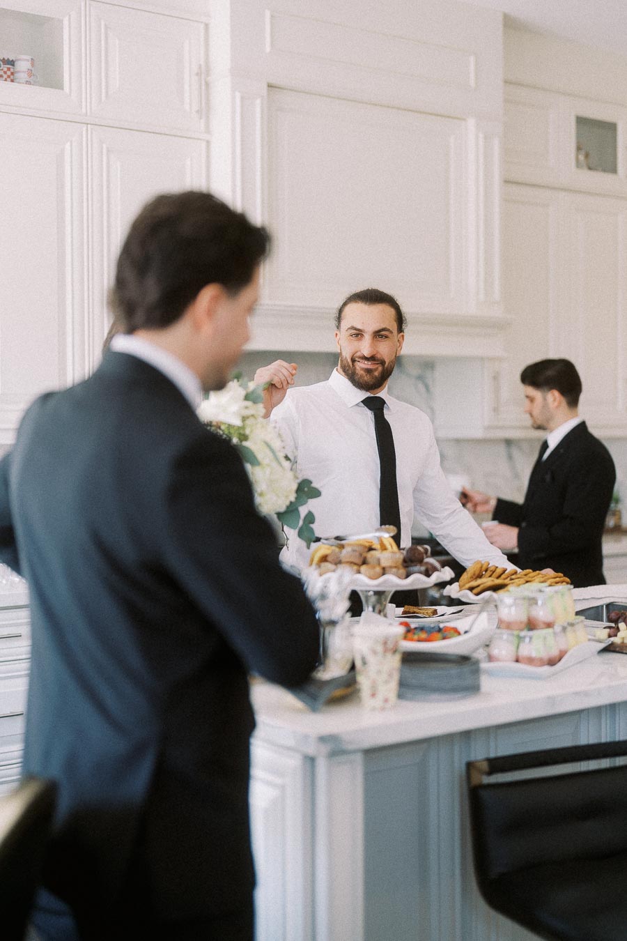 Three men in formal attire enjoying a casual conversation and snacks in a bright, modern kitchen setting. The focus is on a man smiling near a counter laden with pastries and beverages.