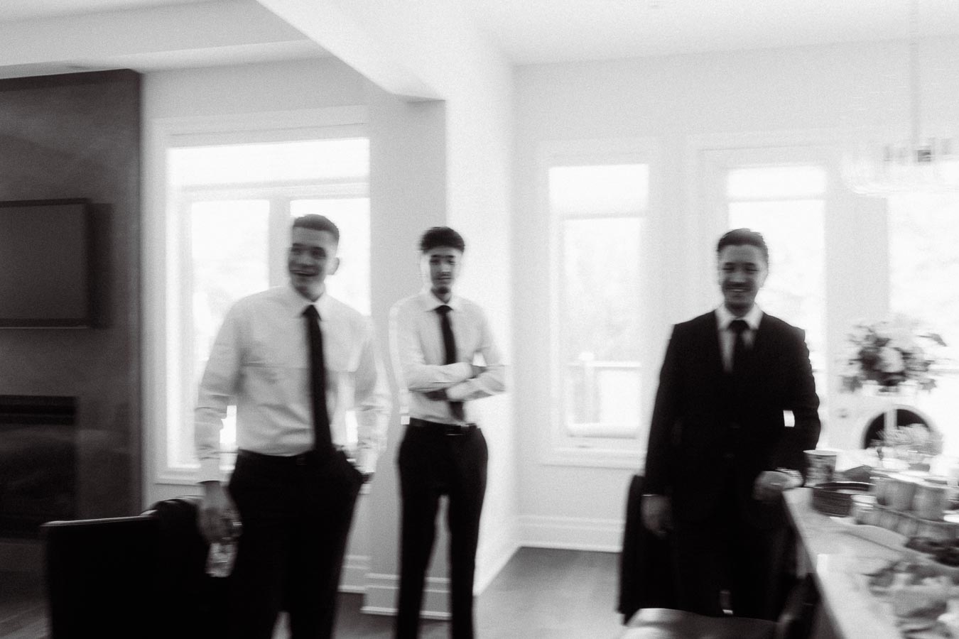 Blurred black and white photo of three men in formal attire standing indoors near large windows, smiling and engaging in conversation.