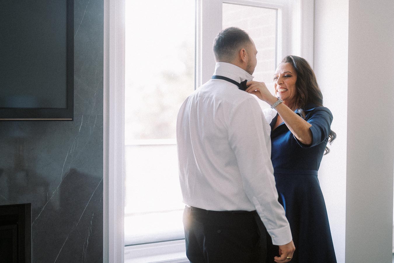 A woman assists a man with his bow tie in a brightly lit room, conveying a sense of warmth and preparation.