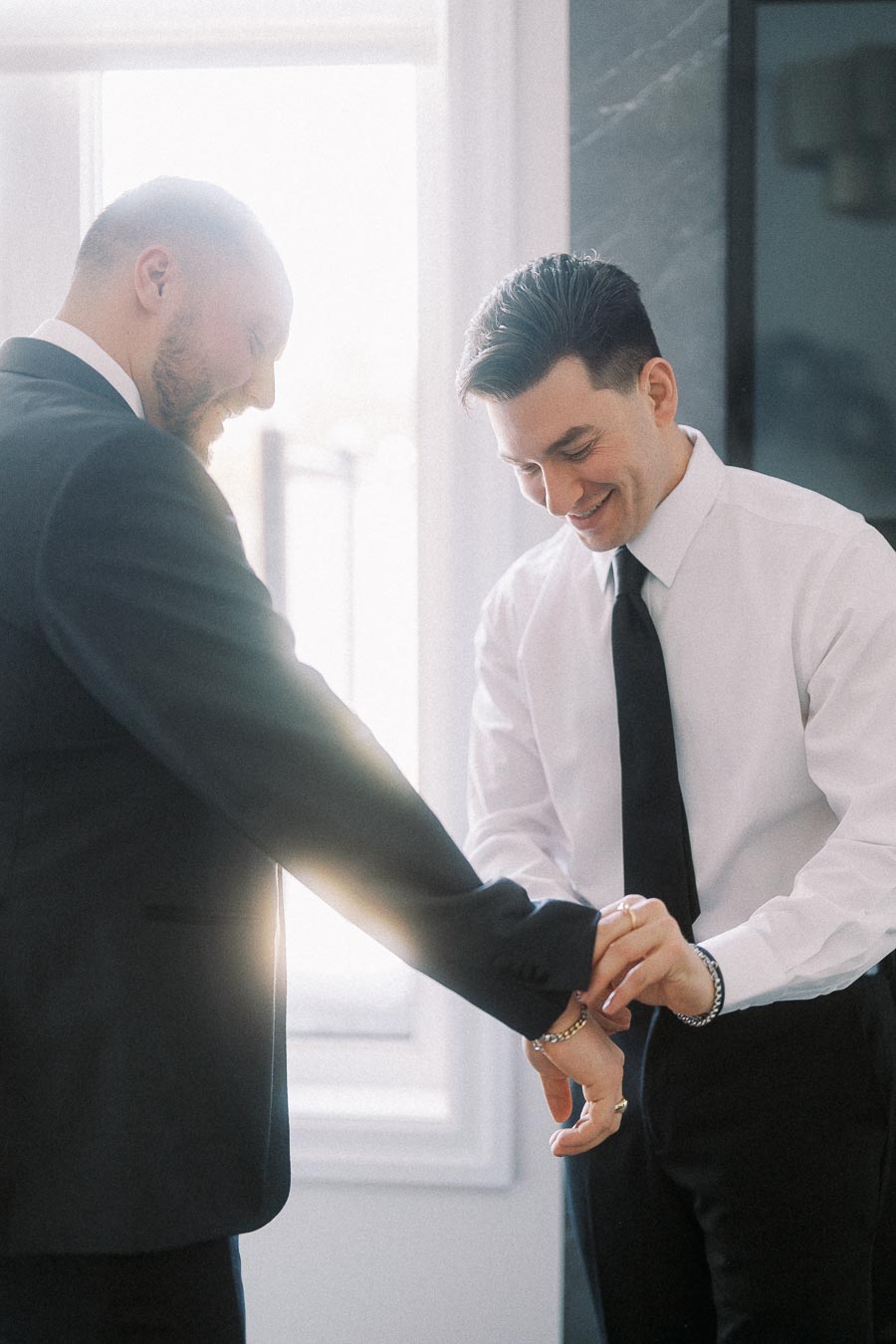 Two men dressed in formal attire smiling while adjusting a suit jacket, backlit by sunlight streaming through a window.