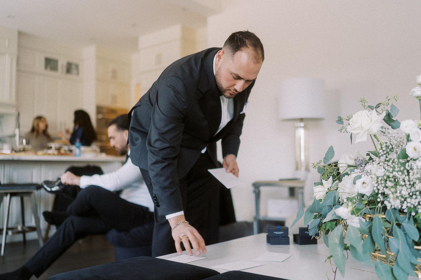 Man in a suit organizing papers in a well-decorated living room with a floral arrangement in the foreground, while people socialize in the background.