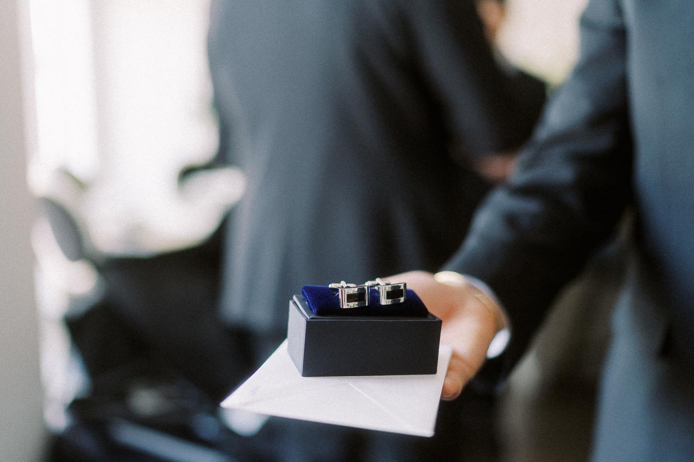 Close-up of elegant silver cufflinks in a black jewelry box, being presented on a white envelope by a person in formal attire.