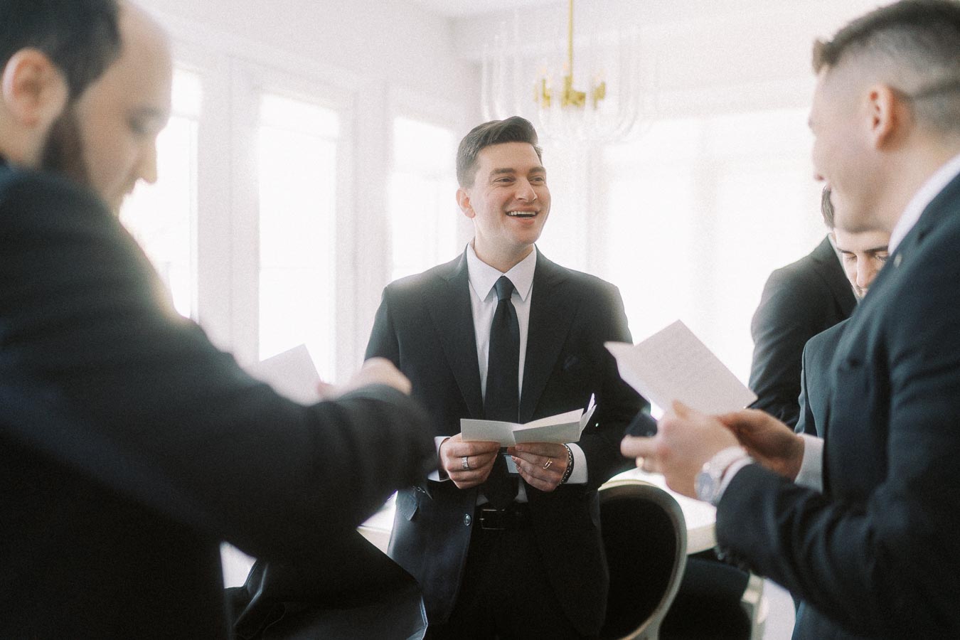 Group of men in suits sharing a light moment and reading notes in a bright room, possibly preparing for an event or celebration.