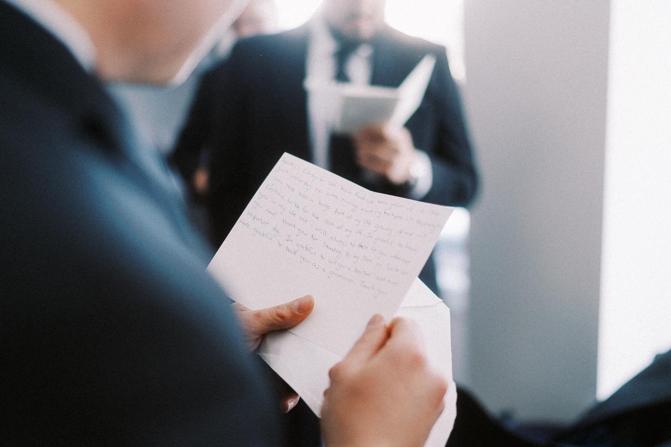 Man in suit reading a handwritten letter at a formal event, with a blurred background of another person in a suit reading a booklet.