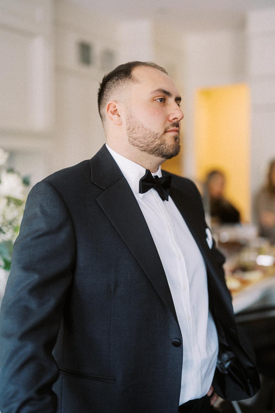Formal event attendee, elegantly dressed in a black tuxedo with a bow tie, stands confidently in a refined indoor setting, hinting at a sophisticated gathering or celebration.