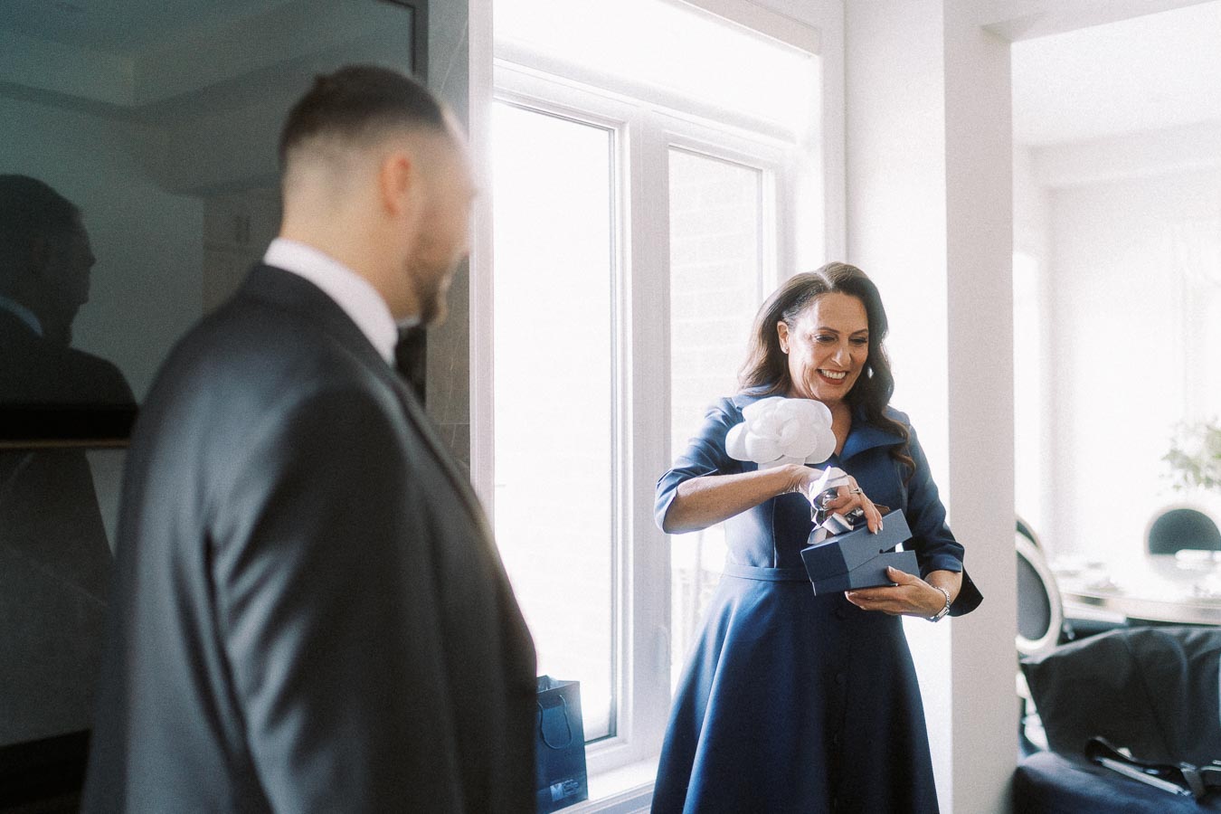 A woman in a blue dress is smiling while opening a gift box with a white flower decoration, standing near a sunlit window. A man in a suit is in the foreground, facing her. The setting appears to be a bright and elegantly furnished room.
