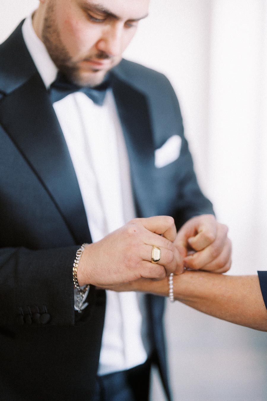 Man in formal black tuxedo adjusting bracelet on another person's wrist.