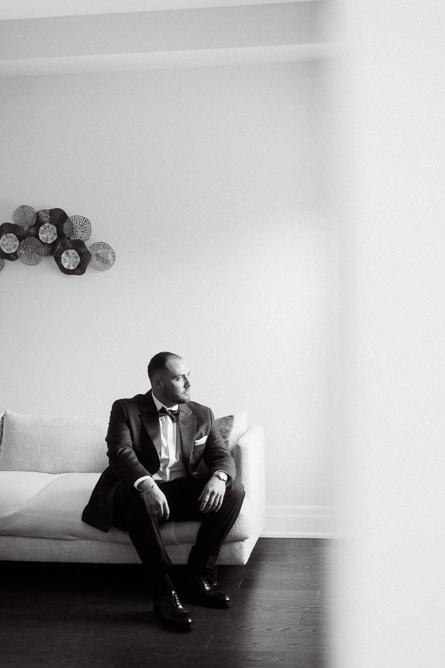 Black and white photo of a man in formal attire sitting on a sofa, looking thoughtfully to the side, with decorative wall art in the background.