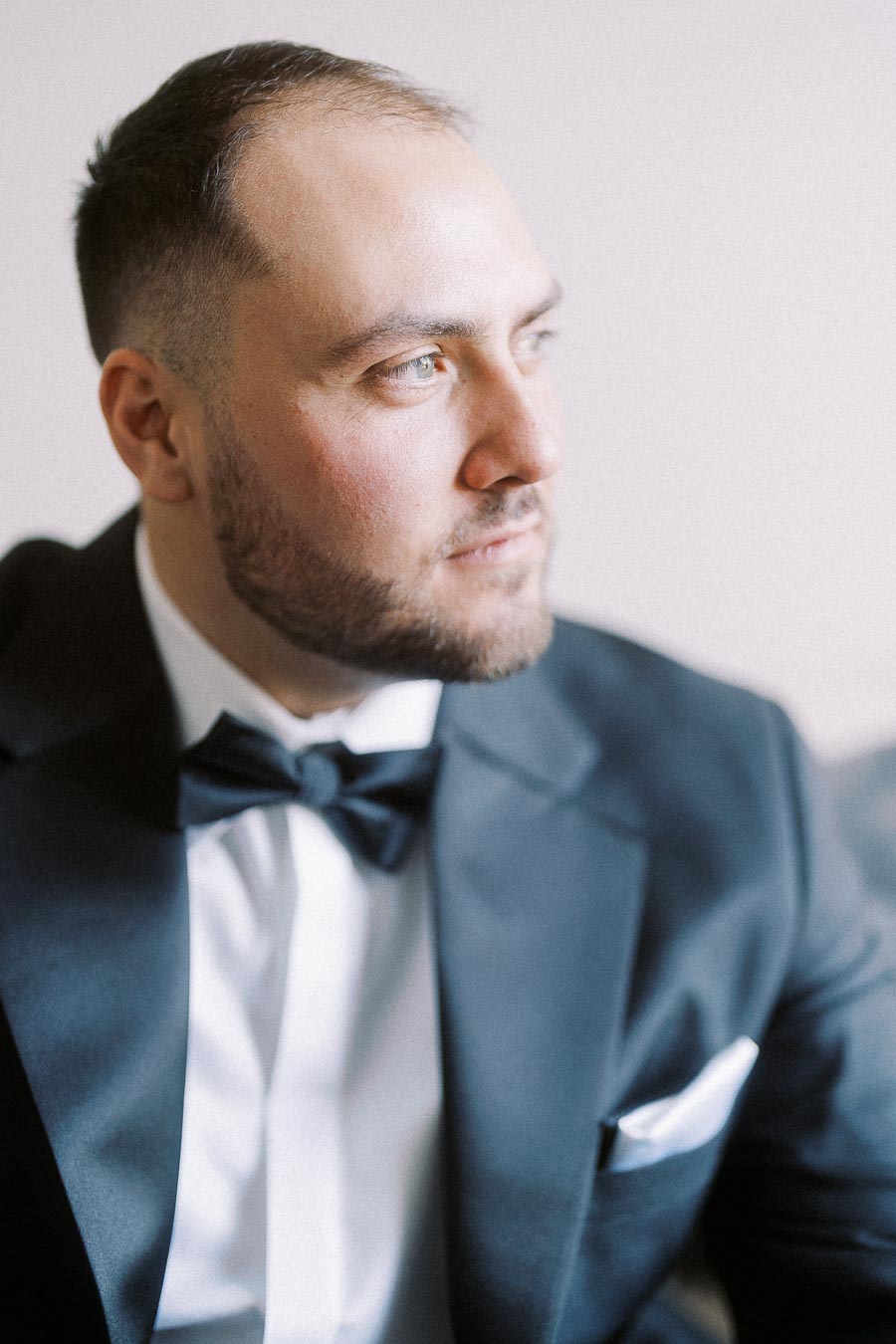 A man in a formal black tuxedo and white shirt looking to the side with a thoughtful expression.