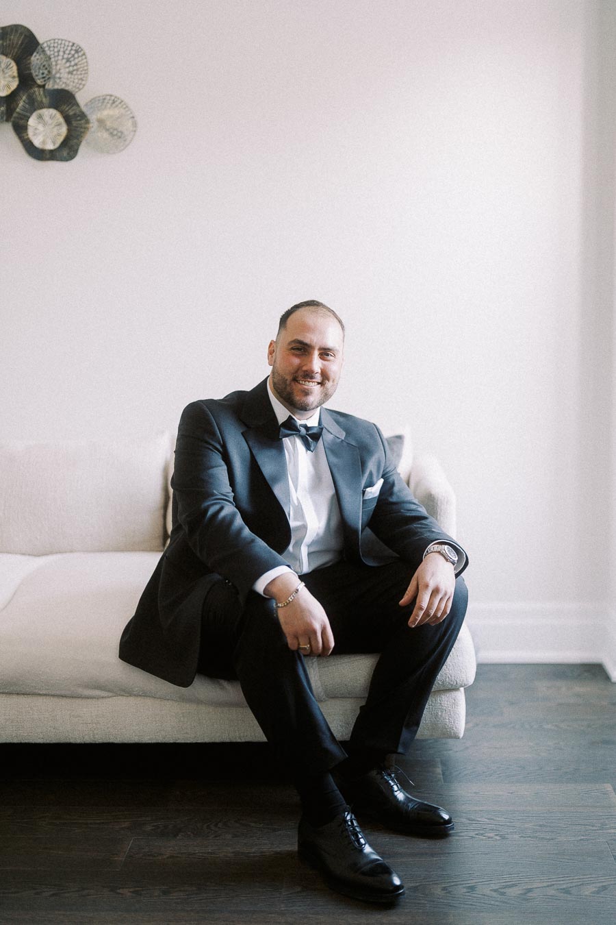 Man in a formal tuxedo sitting on a beige sofa in a well-lit room, smiling confidently, with decorative wall art in the background.