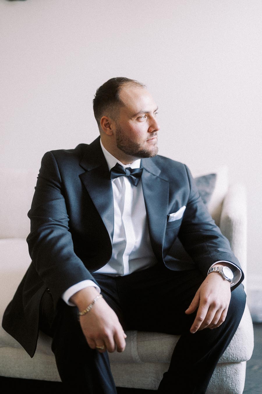 Man in formal suit with bow tie sitting on a light-colored sofa, looking thoughtfully to the side, with subtle lighting highlighting his attire.