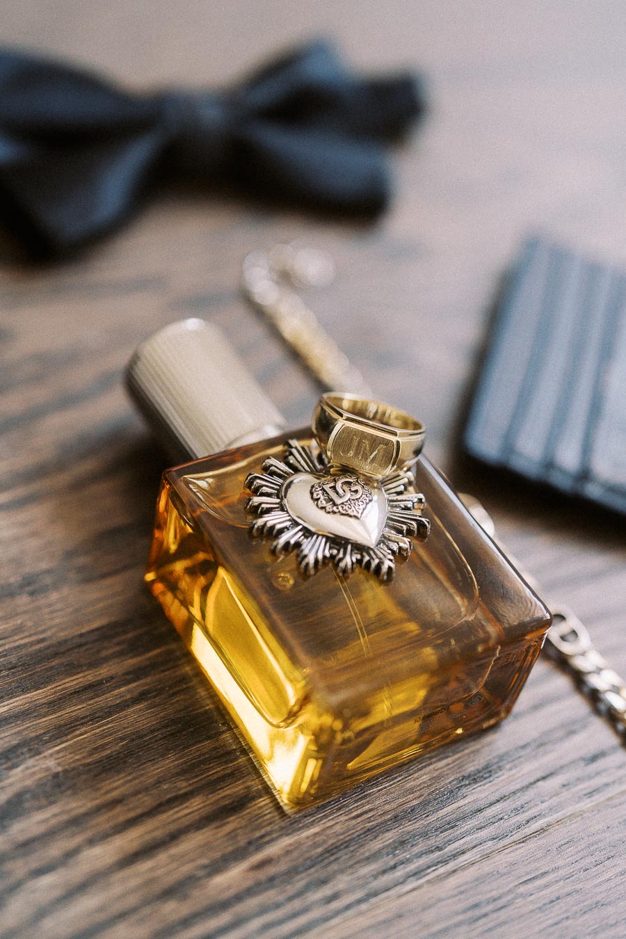 Close-up of a luxurious golden perfume bottle with ornate metallic detailing and a gold ring placed on top, set on a wooden surface. A blurred black bow tie and leather accessory are in the background, highlighting an elegant and sophisticated style.