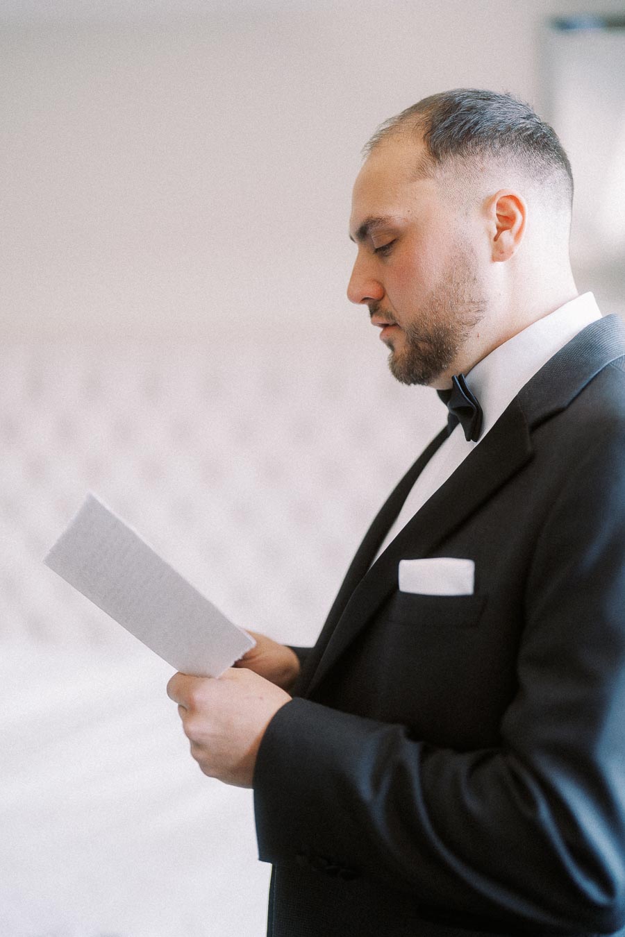 A groom in a black tuxedo reading a letter on his wedding day, standing in a softly lit room.