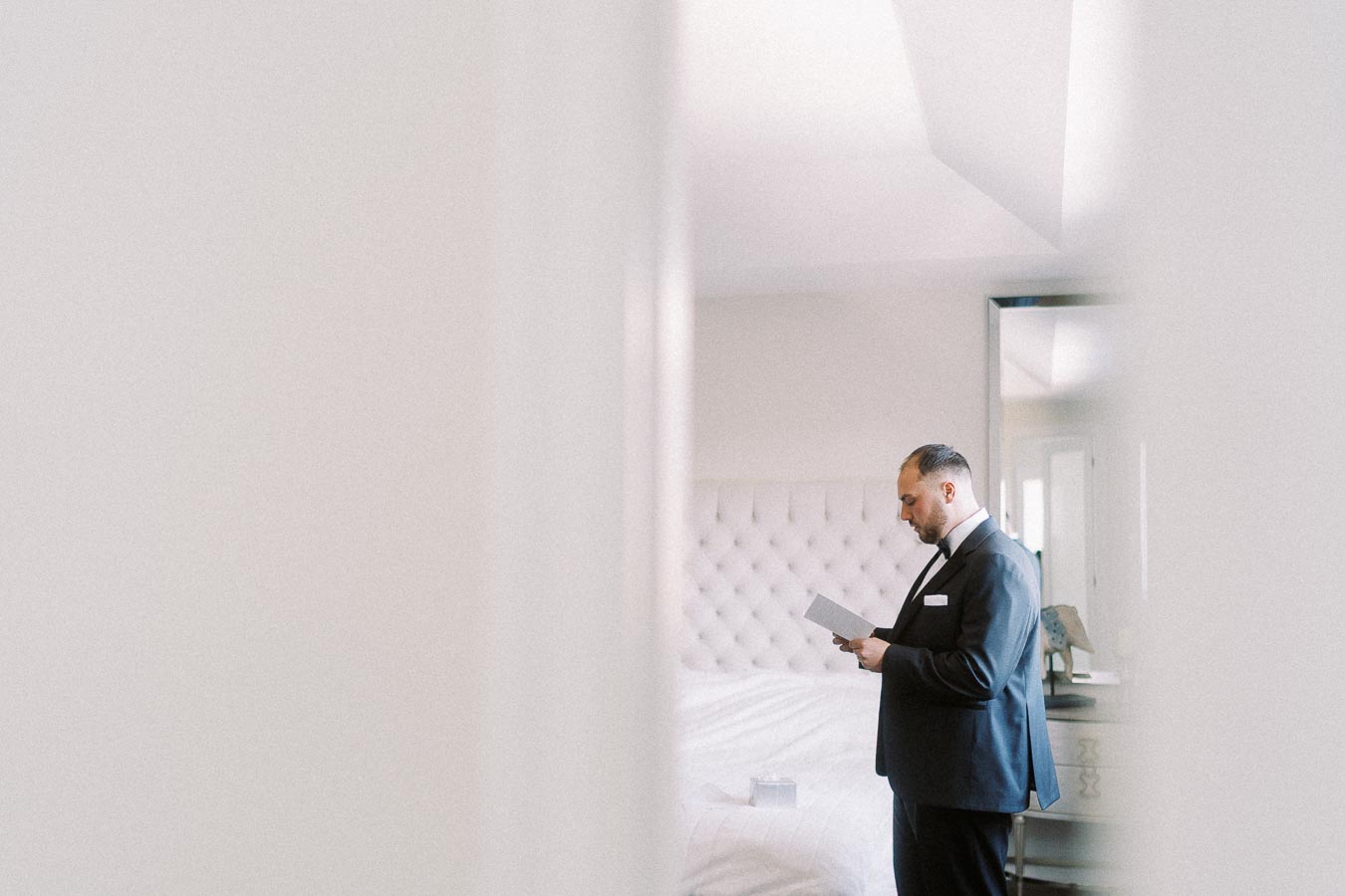 A groom in a suit reads a note in a softly lit modern bedroom, preparing for his wedding day.
