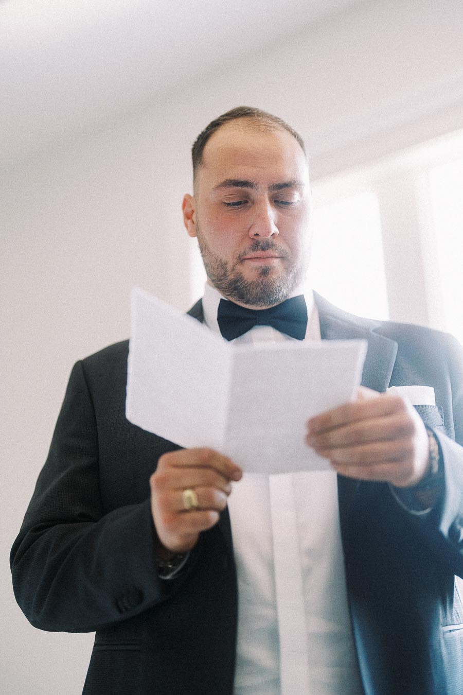 Elegant man in a black tuxedo reading a note, standing in a bright room with natural light streaming through a window.