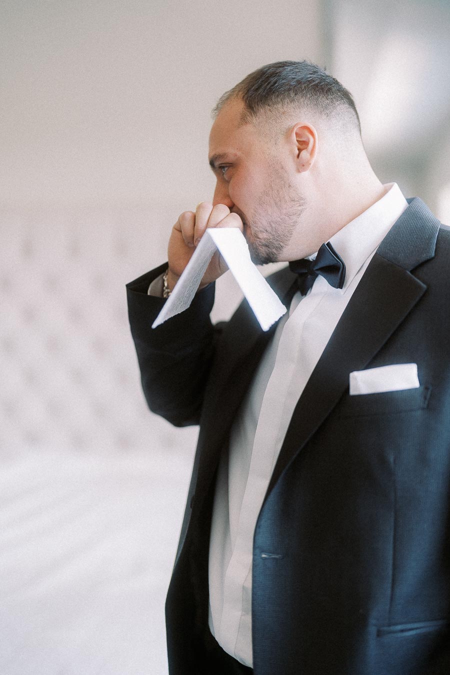 Elegant groom in a black tuxedo holding a folded letter, showcasing emotion and anticipation before a wedding ceremony.