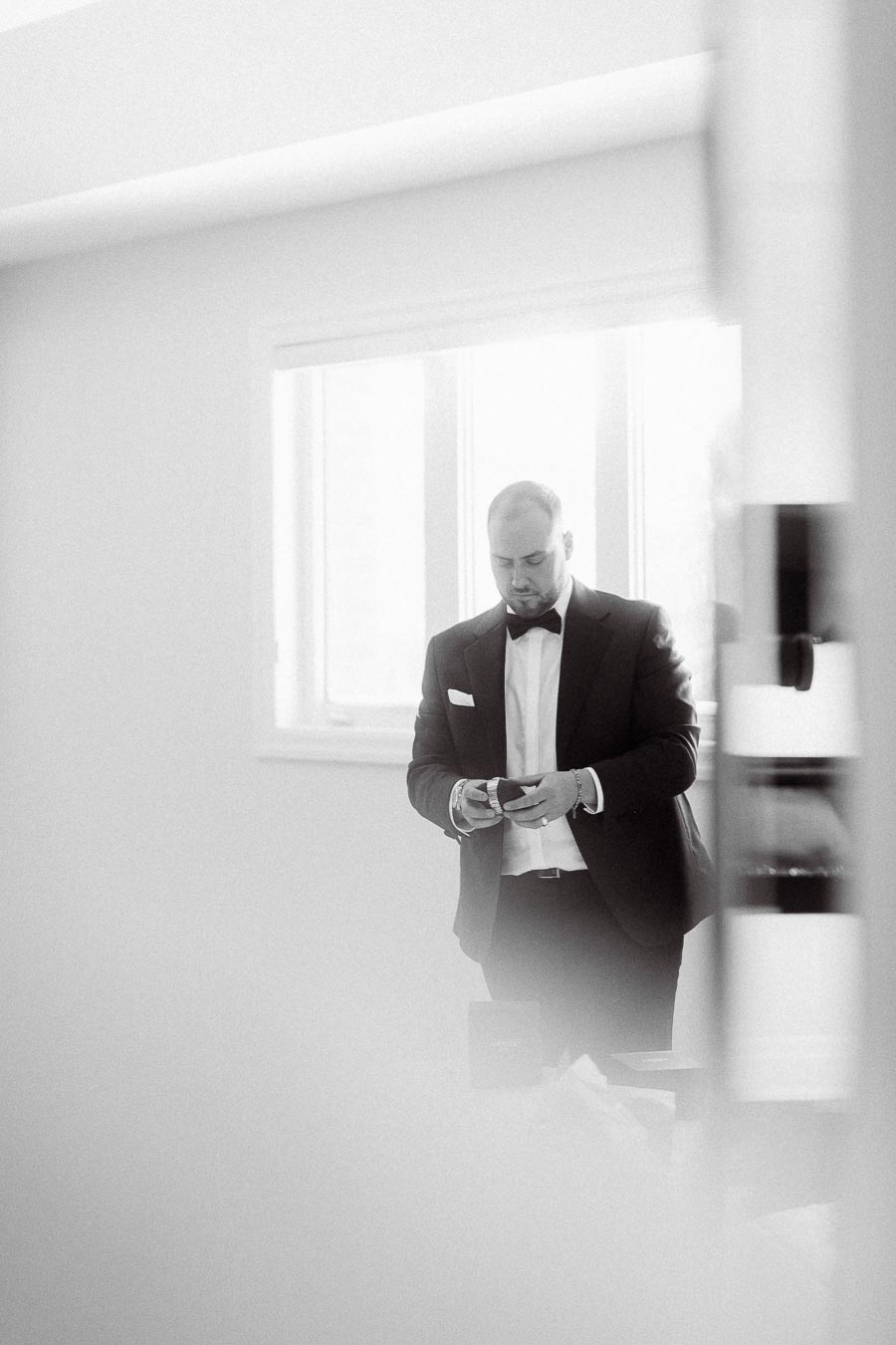 Black and white photo of a man in formal attire adjusting his bow tie in a softly lit room, conveying elegance and preparation.