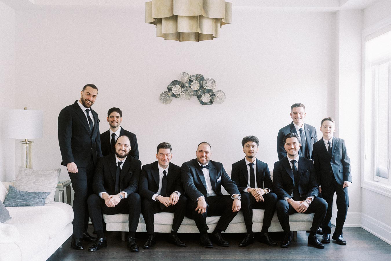 Group of men in suits posing for a formal photograph in a stylish, modern living room with a decorative wall piece and chandelier in the background.