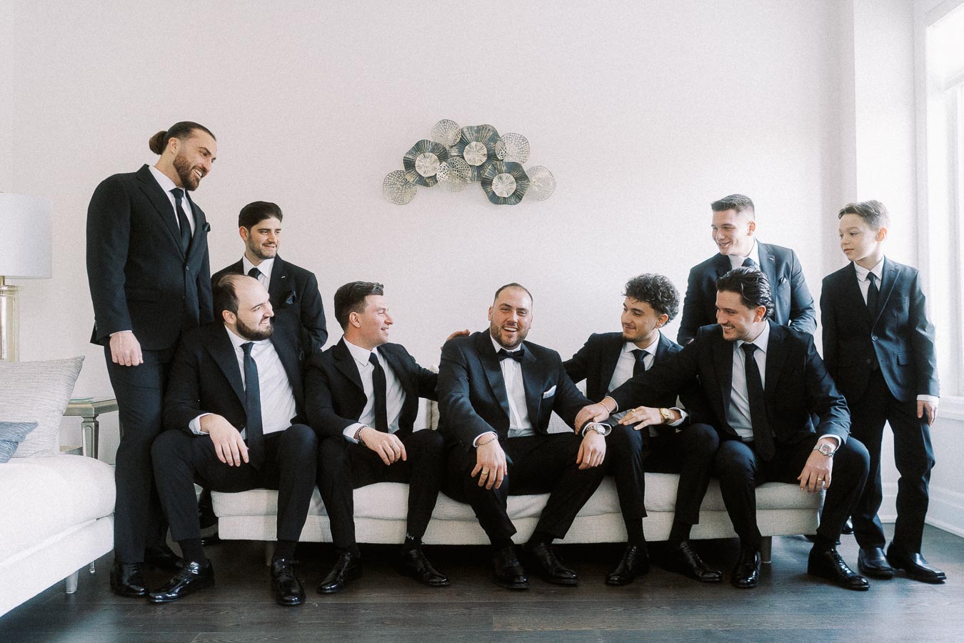 Groom with groomsmen laughing and sitting on a white sofa, wearing formal black suits and ties, in a bright room.