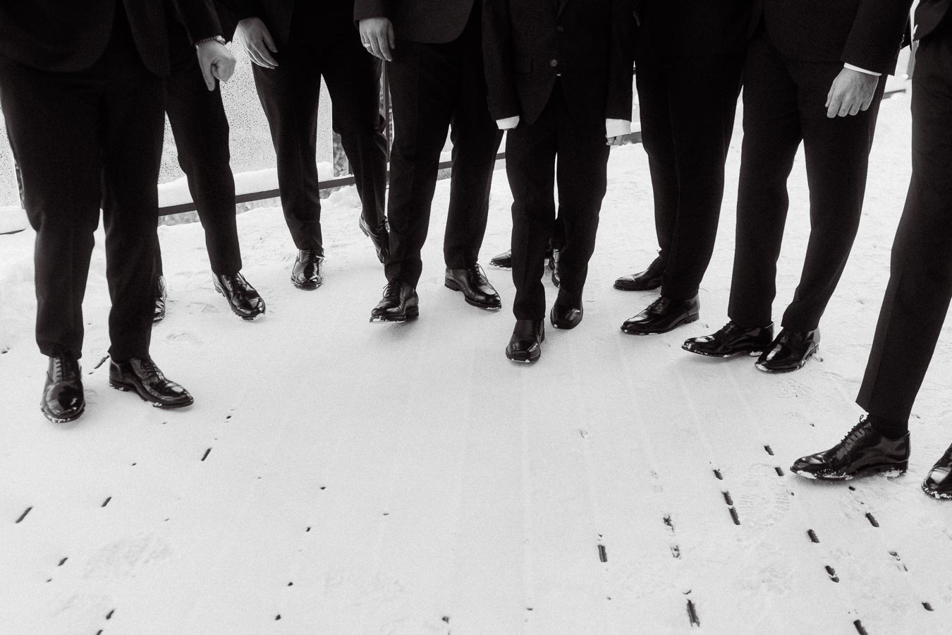 Group of people in formal attire standing on a snowy surface, focusing on polished shoes and suits. Black and white photo highlighting the elegance and sophistication of a wedding or formal event.