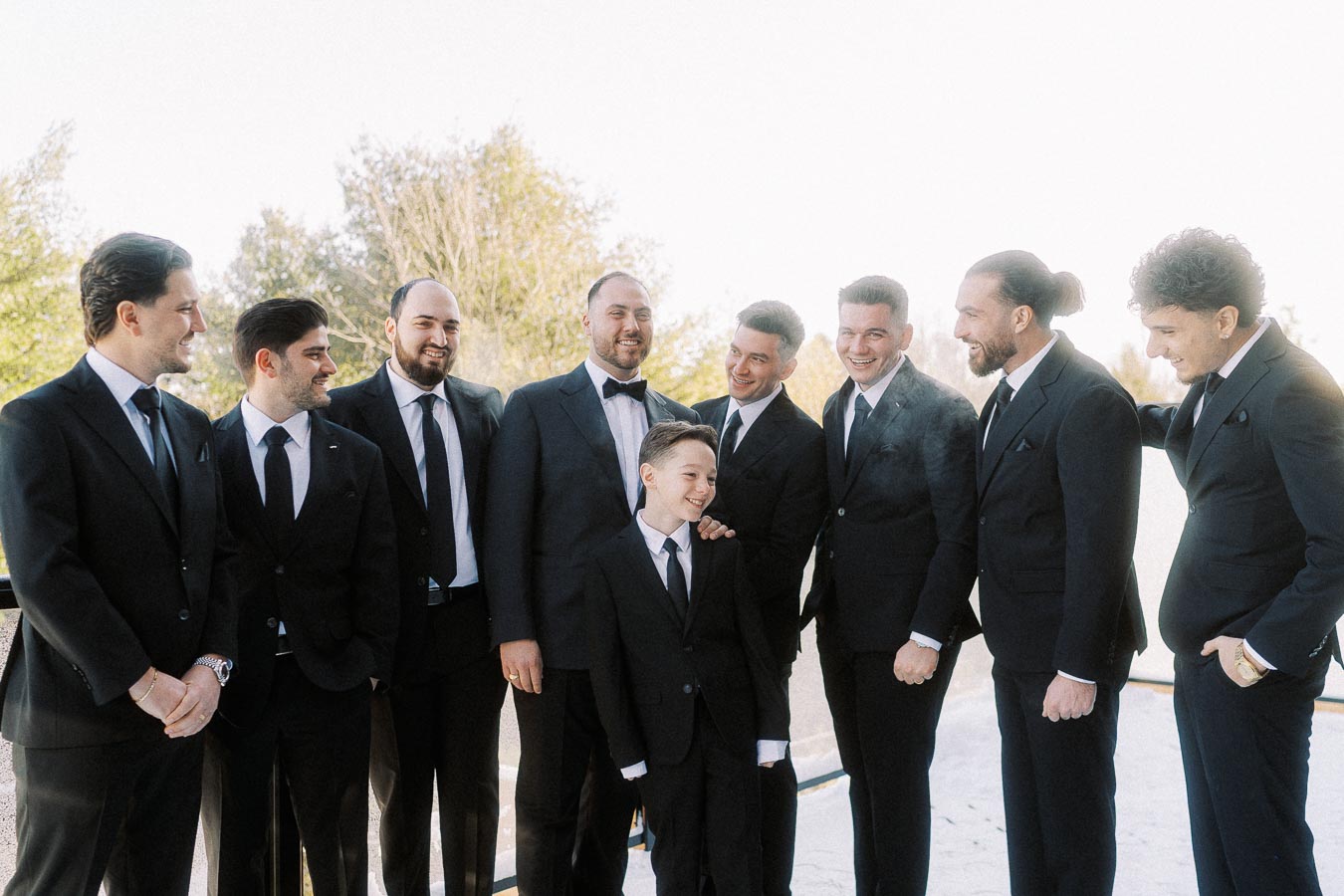Group of groomsmen and a young boy in suits and tuxedos laughing together outdoors, capturing a joyful wedding moment.