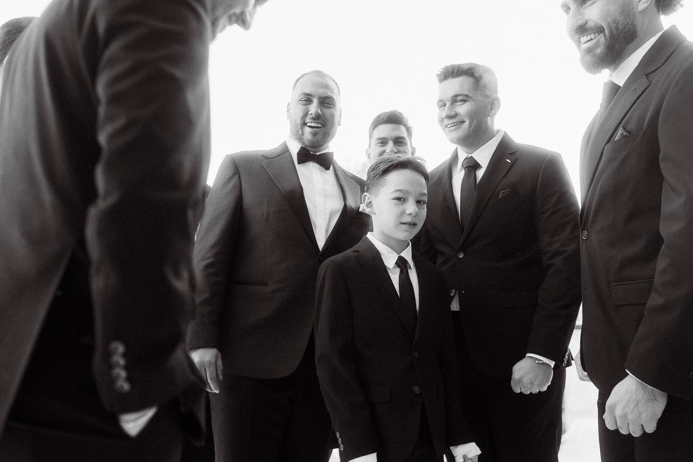 A group of men and a young boy in formal suits smiling and interacting, possibly at a wedding or formal event, conveying a sense of celebration and camaraderie.