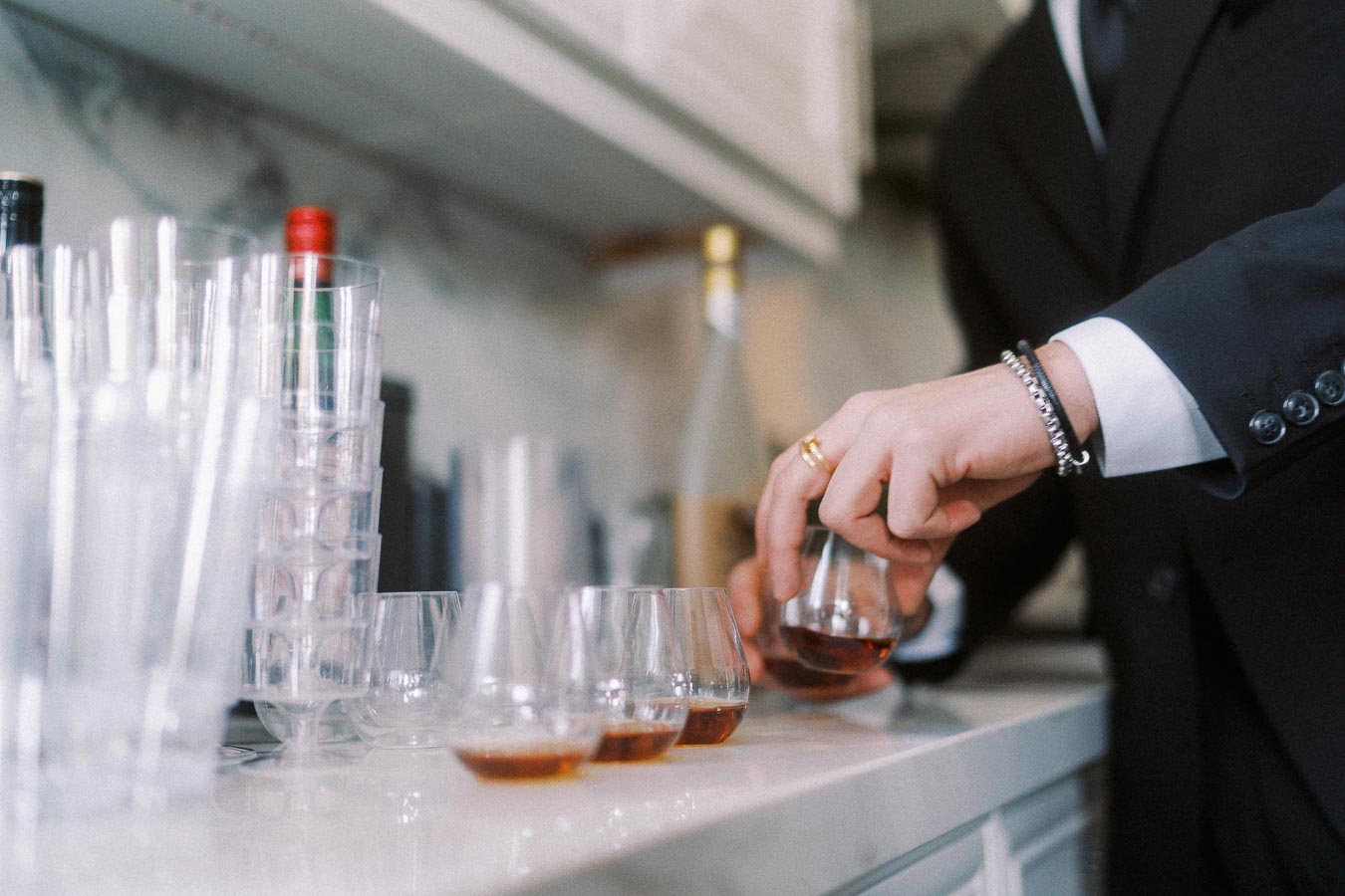 A person in a formal suit pouring whiskey into glasses on a bar counter, surrounded by stacked empty cups and bottles in the background.
