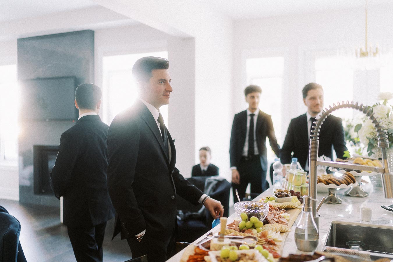 Group of well-dressed men in suits enjoying a gathering around a kitchen island with a variety of appetizers, fruits, and drinks in a bright, modern home interior.