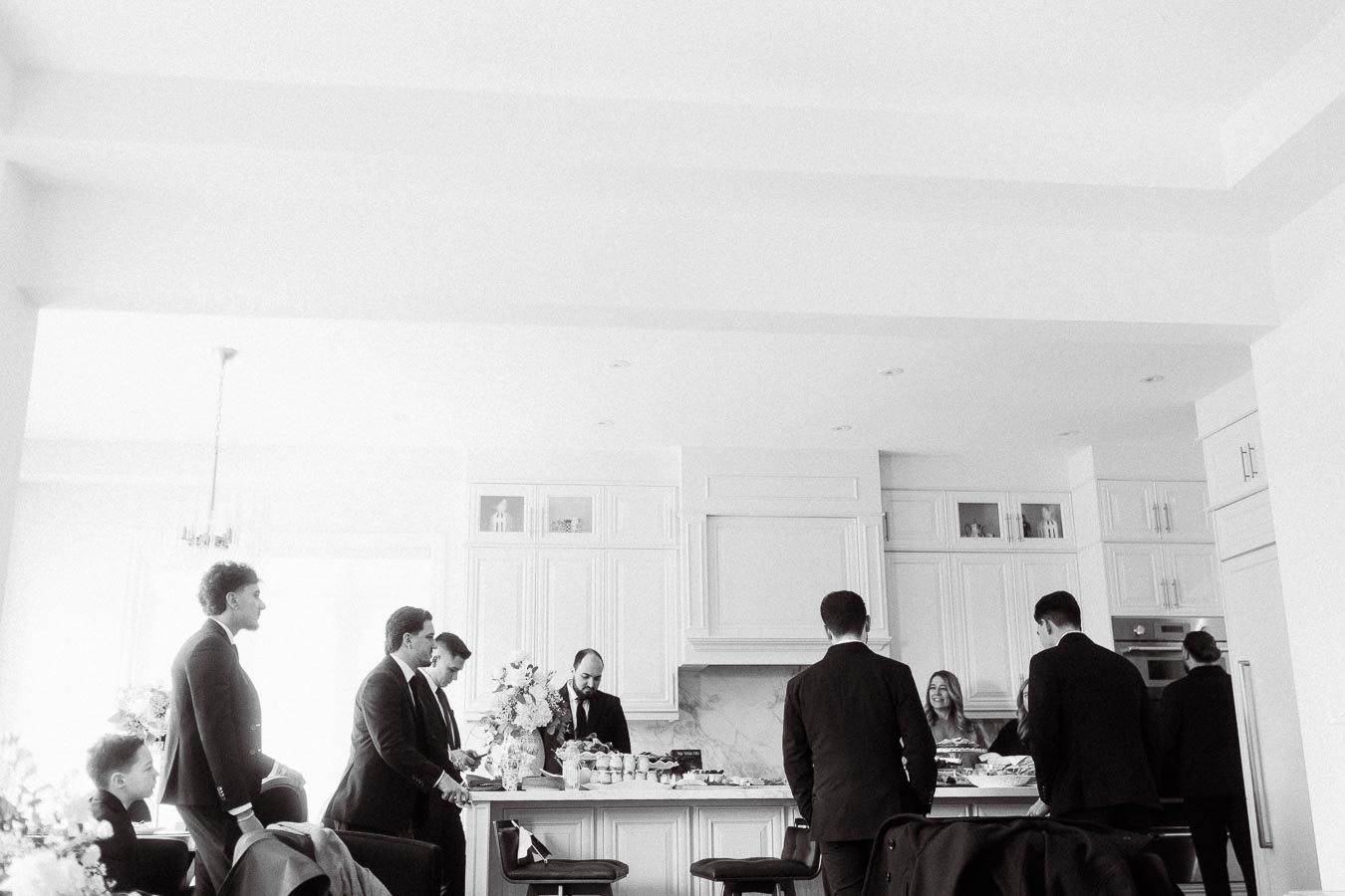 Black and white image of a group of people dressed in formal attire, gathered around a kitchen island with food and drinks, in a bright, modern kitchen setting.