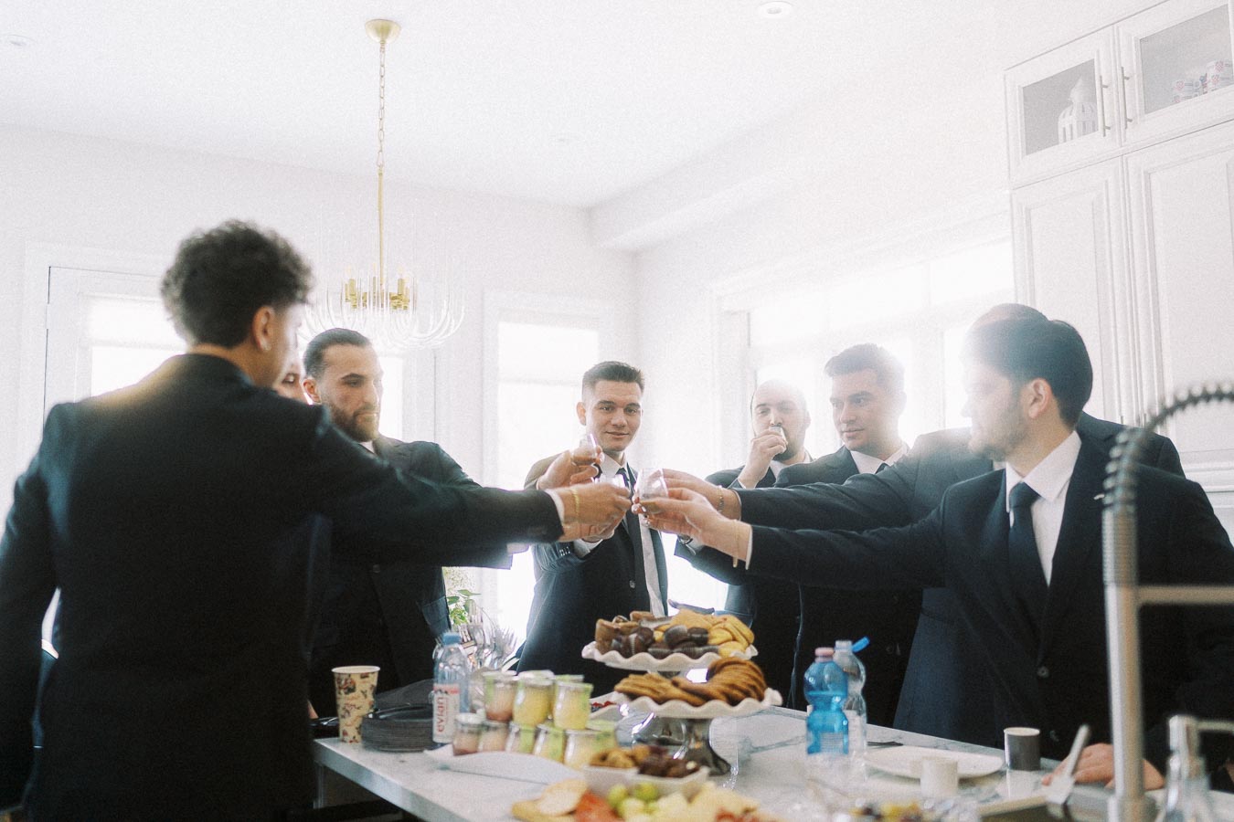 Group of men in suits toasting glasses in a bright kitchen setting, with a variety of desserts and beverages on the countertop, creating a celebratory atmosphere.