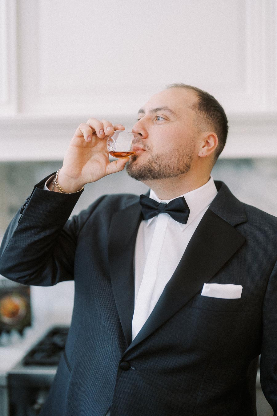 A man in a formal black suit and bow tie drinks from a glass in an elegant setting.