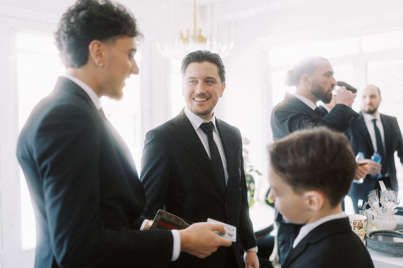 A group of well-dressed men in suits socializing at an indoor event, with one man smiling and holding a card, creating a lively and formal atmosphere.