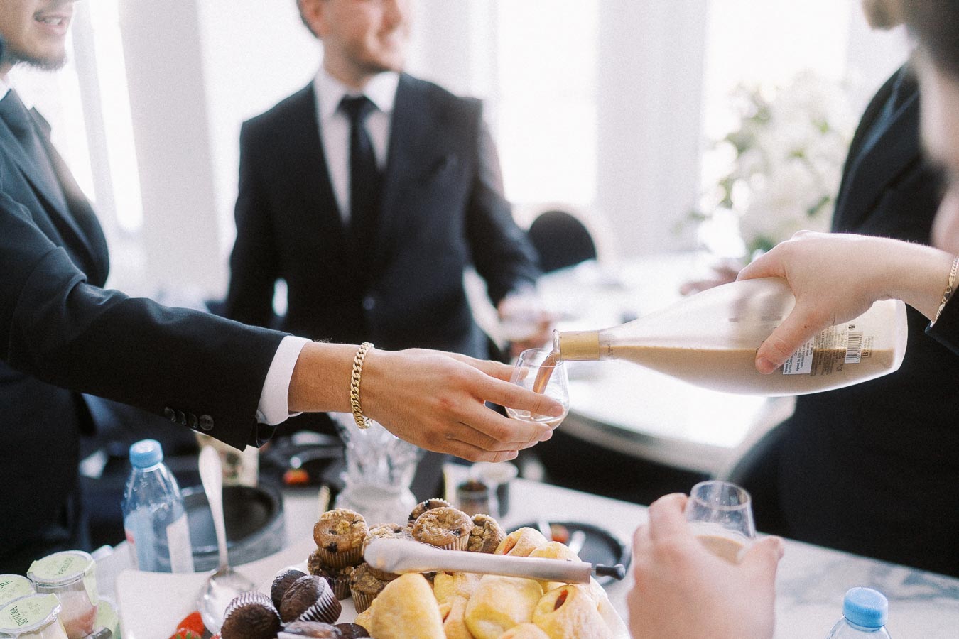 Business professionals in suits enjoying a catered event with an assortment of pastries and drinks, featuring a close-up of a hand pouring a beverage into a glass, creating a sophisticated networking atmosphere.