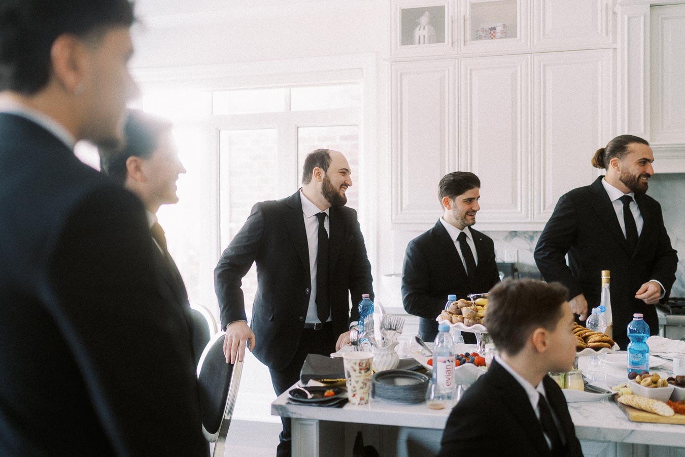 A group of men in suits gather around a kitchen island, enjoying snacks and drinks, conveying a festive and celebratory atmosphere.