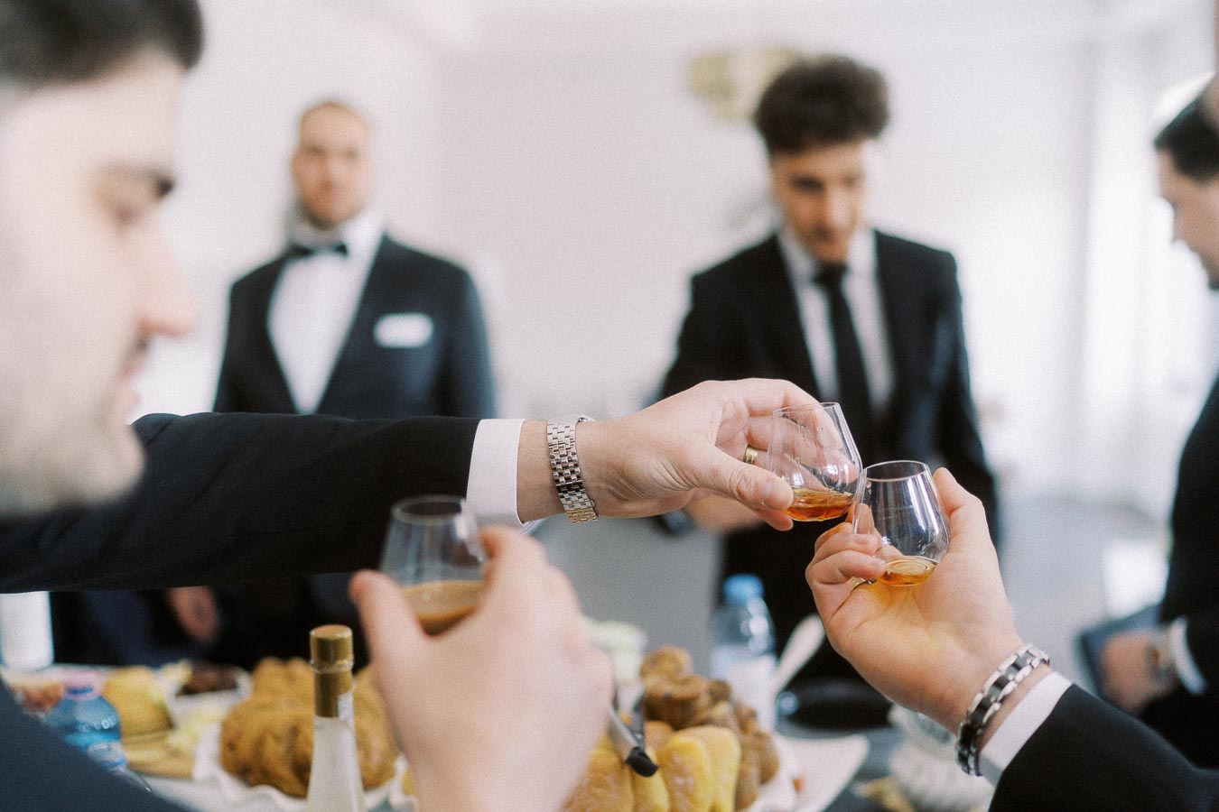 Group of men in suits toasting with glasses of whiskey during a formal gathering, with appetizers and a bright, elegant setting in the background.