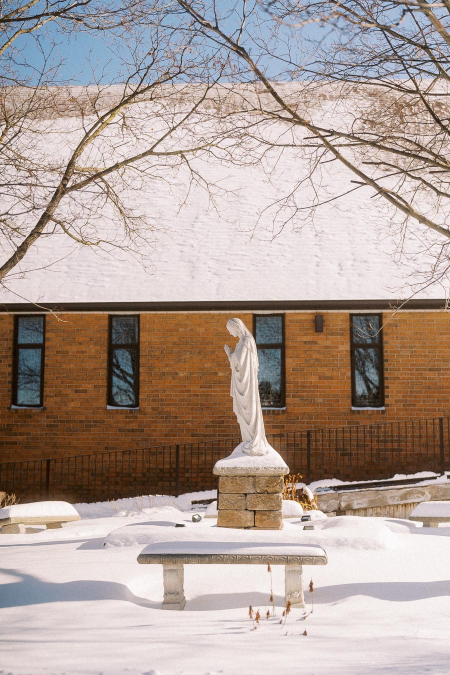 Statue of the Virgin Mary surrounded by snow in front of a brick church building, with bare tree branches and a clear blue sky.