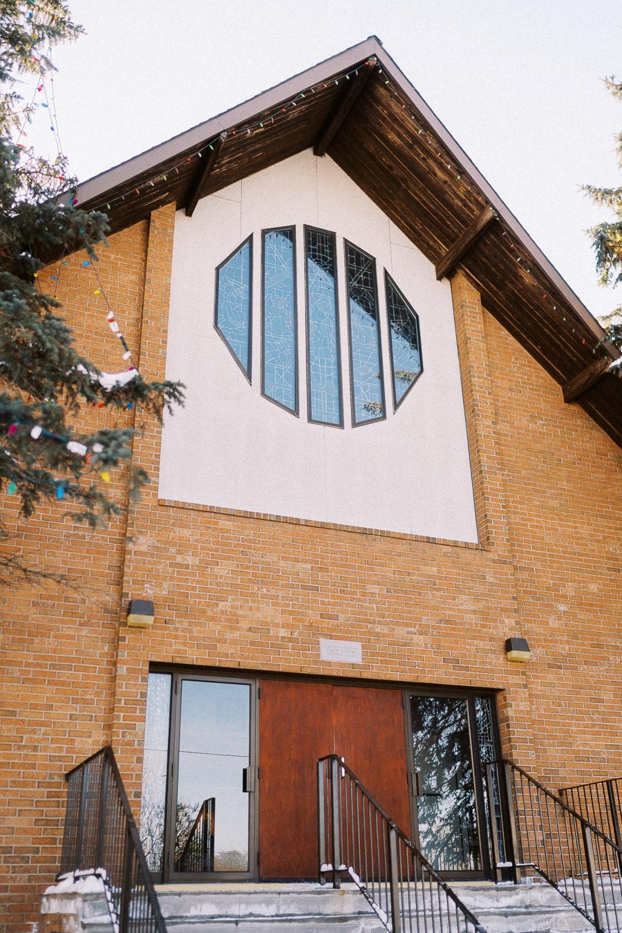 Front view of a brick church with decorative stained glass windows and wooden doors, adorned with overhead lights and surrounded by trees on a winter day.