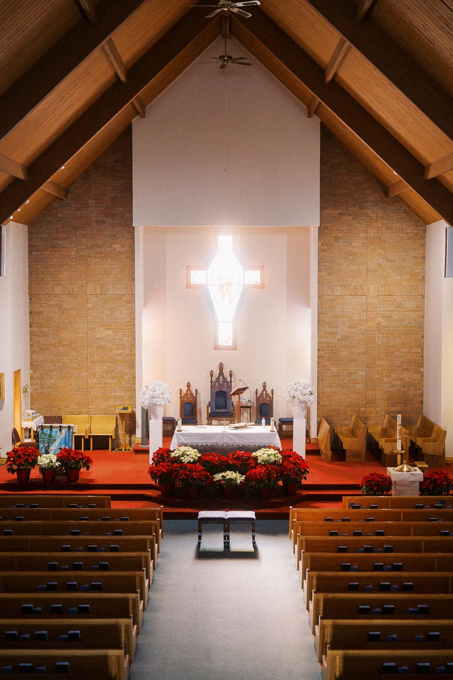 Interior of a church featuring a wooden vaulted ceiling, central altar adorned with red and white floral arrangements, wooden pews, and a bright, illuminated cross on the wall.