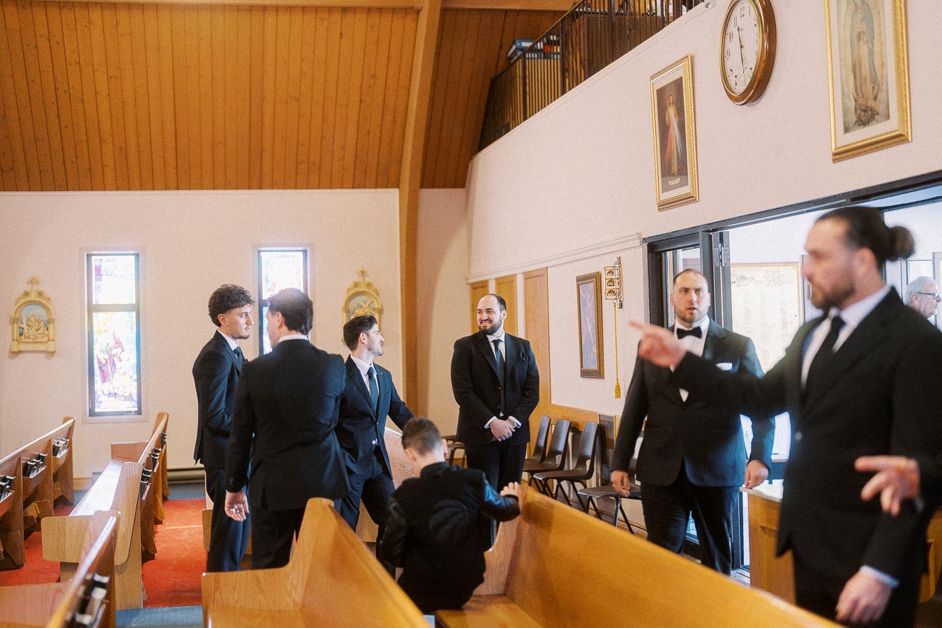 A group of men in formal suits, likely groomsmen, gather and converse inside a church with wooden pews and stained glass windows.