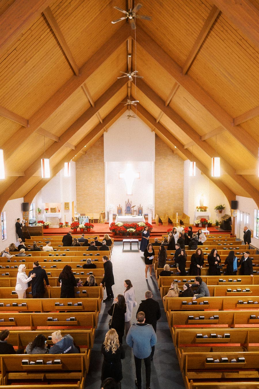 A congregation gathering inside a church with wooden pews and high ceilings, people socializing in the aisle near the altar decorated with red and white flowers.