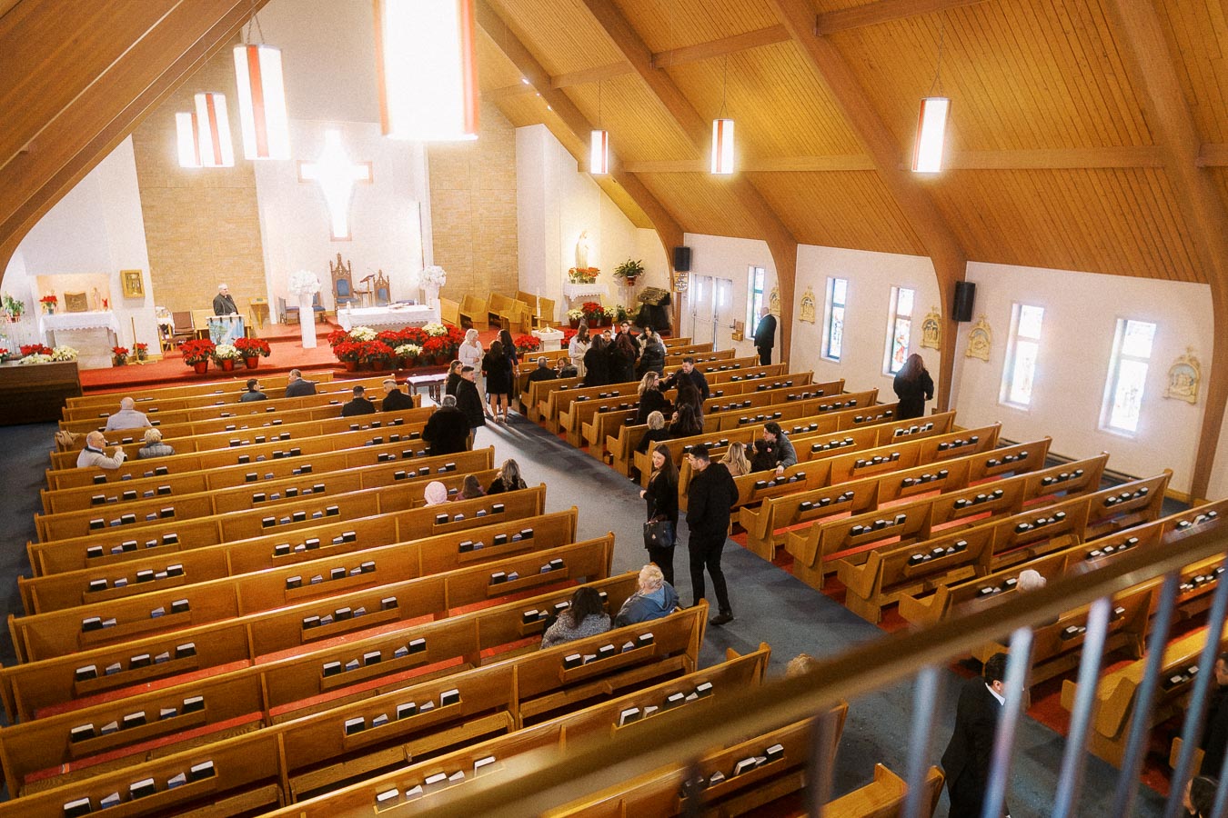 Interior view of a spacious, well-lit church with wooden pews, attendees gathering, and a prominent altar adorned with floral arrangements, conveying a serene and welcoming atmosphere.