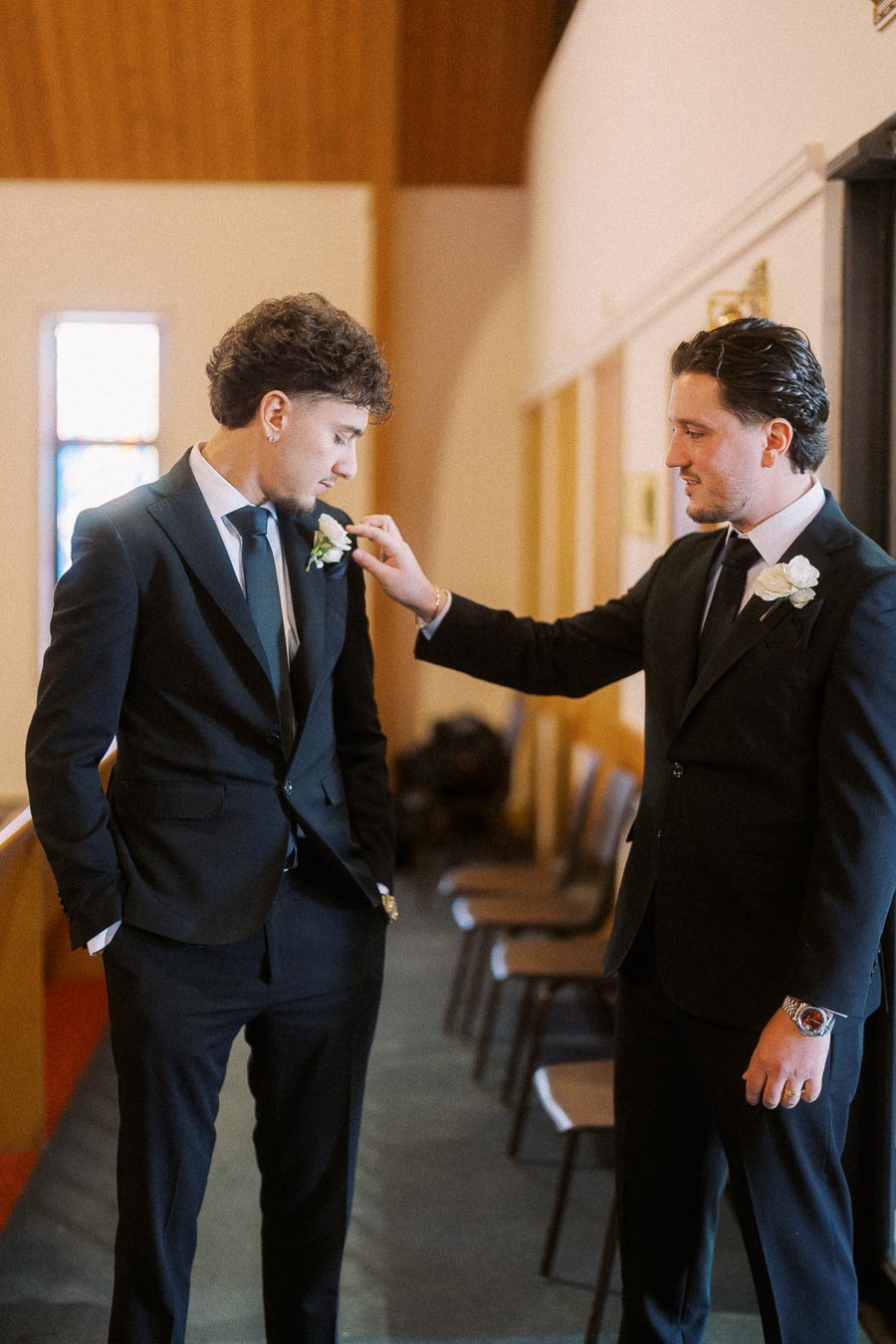 Two men in suits inside a room, one adjusting the other's boutonniere, suggesting preparation for a formal event such as a wedding.