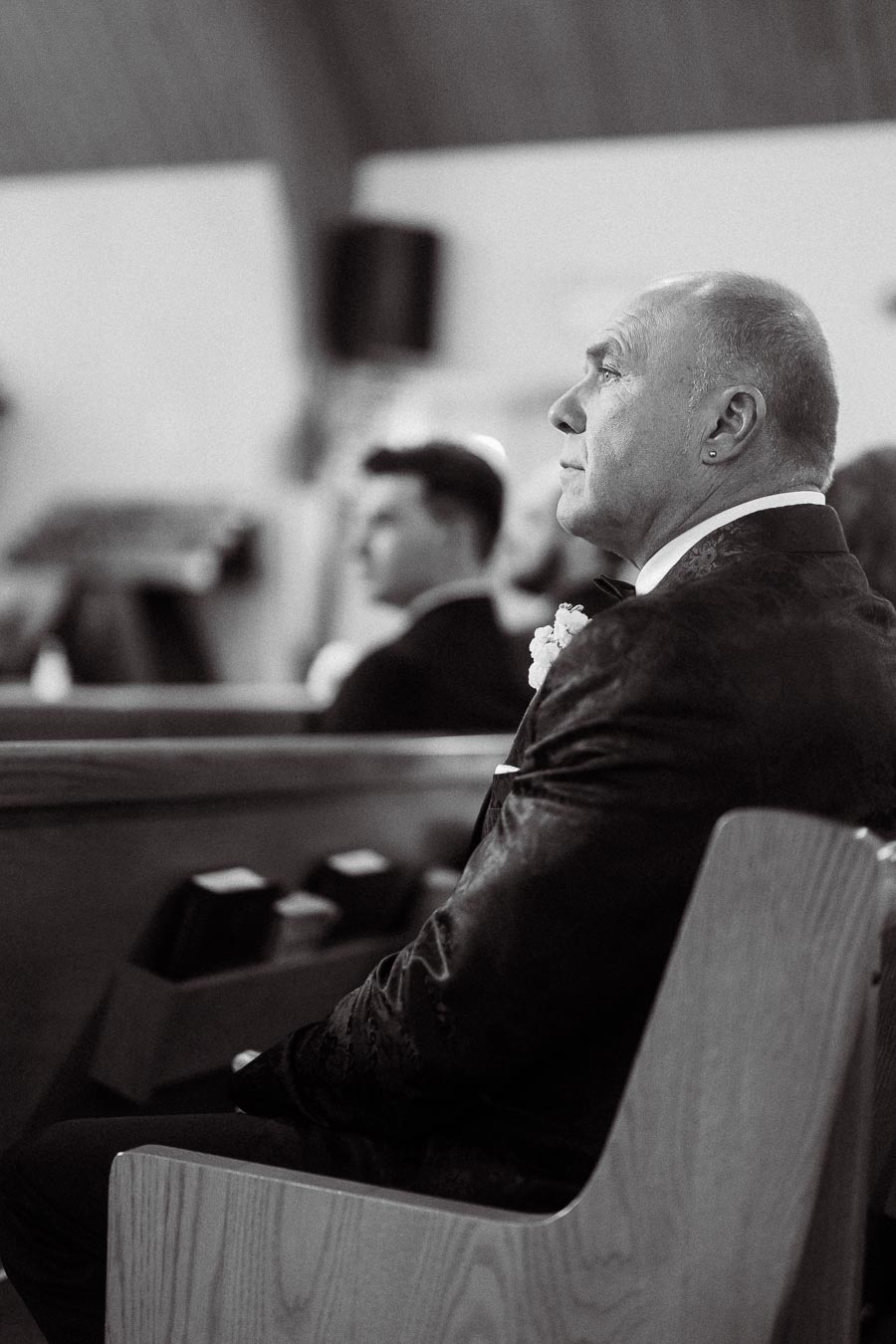 Elderly man in formal attire sitting in a church pew, attentively watching a ceremony, with blurred background and ambient lighting.