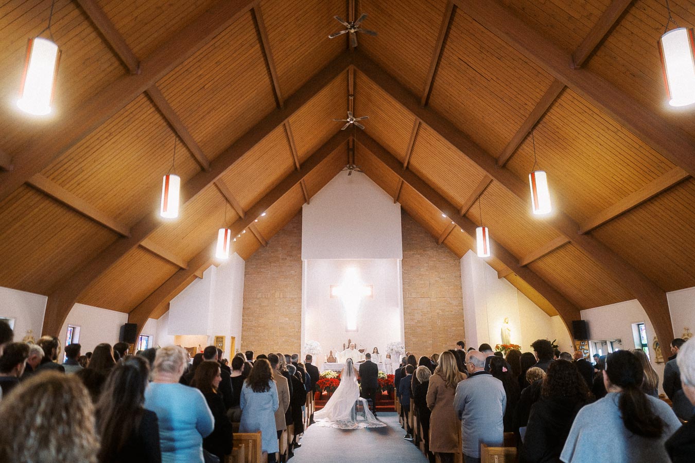 A congregation attending a wedding ceremony in a beautifully lit church with a wooden vaulted ceiling and a prominent cross at the altar.