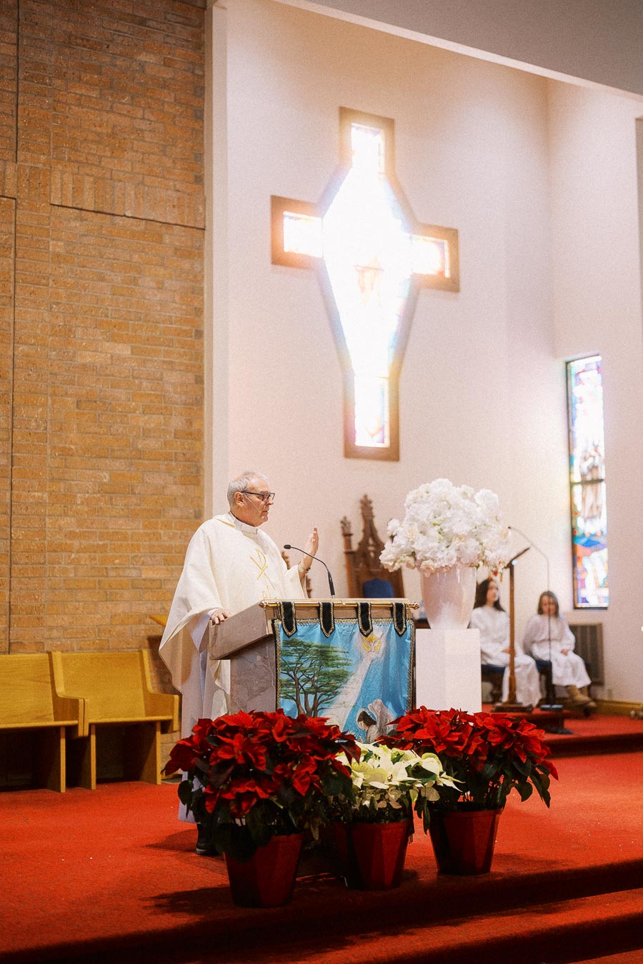 Priest delivering a sermon from a pulpit in a church with a large cross-shaped stained glass window behind, surrounded by red and white floral arrangements and seated choir members.