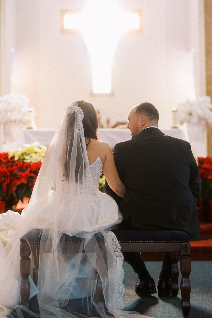 Rear view of bride and groom sitting in a church during wedding ceremony, with soft lighting and a large illuminated cross in the background.