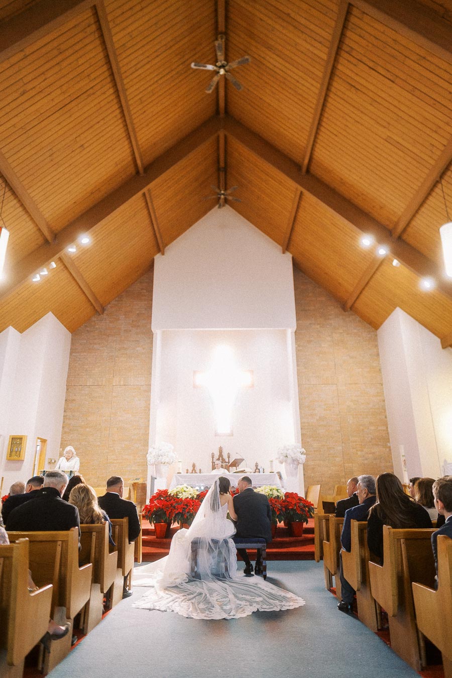 A bride and groom kneeling at the altar during a church wedding ceremony, surrounded by seated guests and festive decorations; a large cross is illuminated in the background.