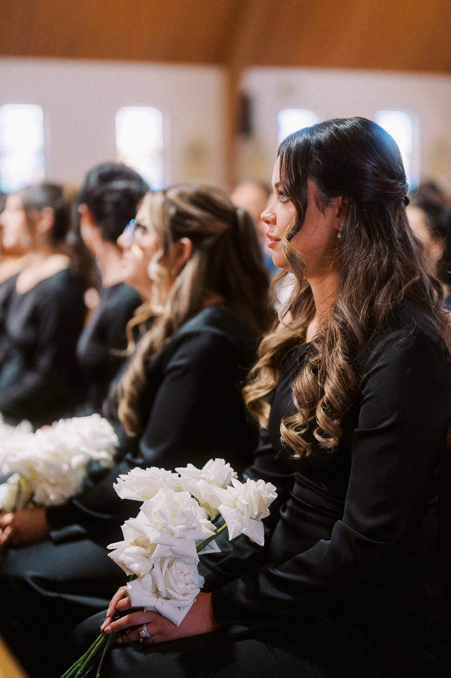 Bridesmaids in elegant black dresses holding white rose bouquets, seated in a row during a wedding ceremony.