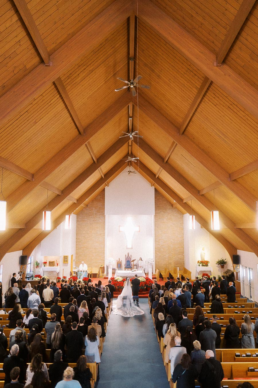 Wedding ceremony in a beautiful wooden-ceiling church filled with guests, bride walking down the aisle toward the altar.