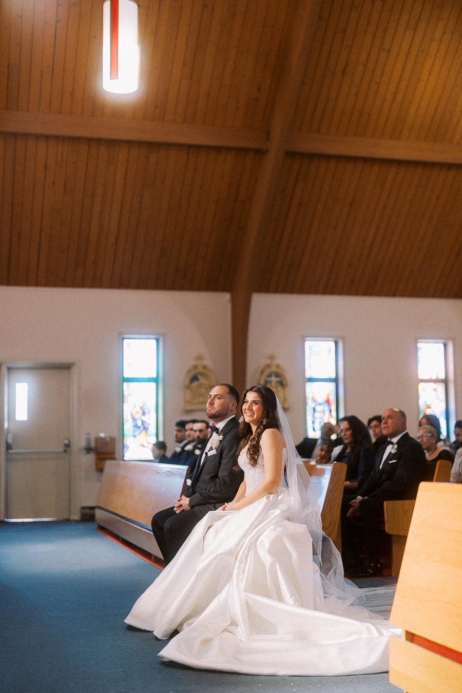 Bride and groom seated during a wedding ceremony in a beautifully lit church with stained glass windows and wooden pews.